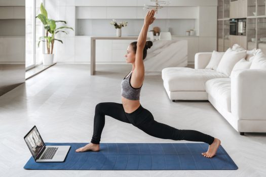 Beautiful young woman in sports clothing practicing yoga at home, watching online tutorial, repeating asans, standing in Warrior one pose, doing Virabhadrasana exercise. Fit, healthy, wellness concept